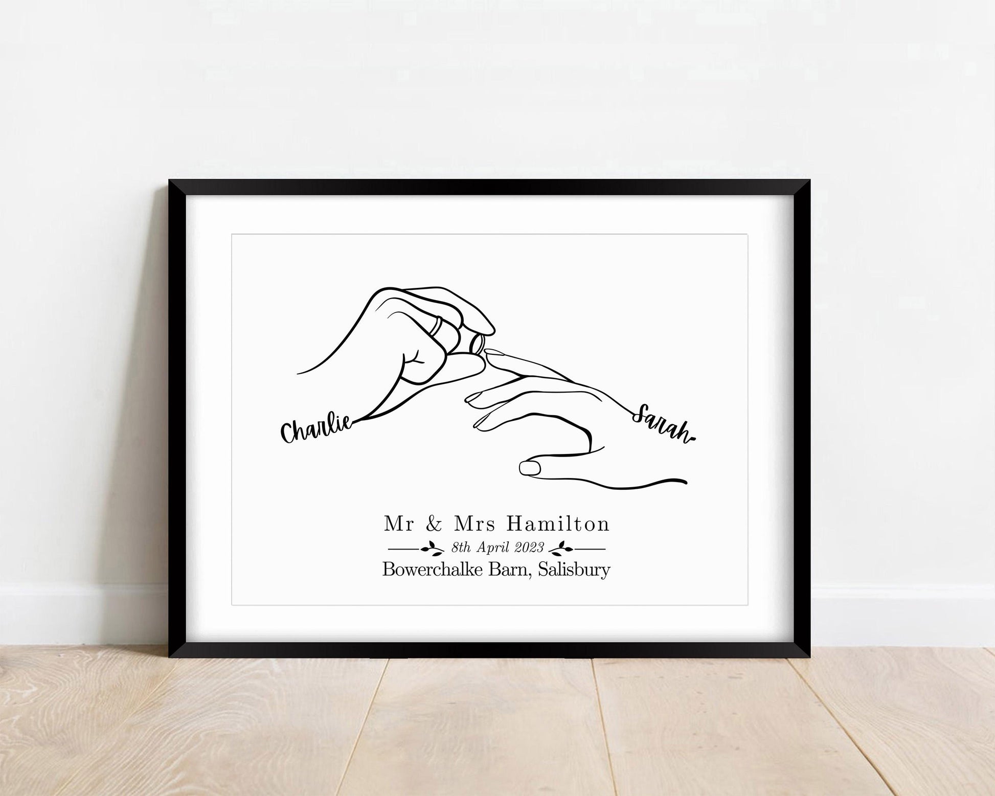 Framed custom wedding print featuring minimalist line art of two hands, with one person placing a wedding ring on the other. The hands are labeled with the names.