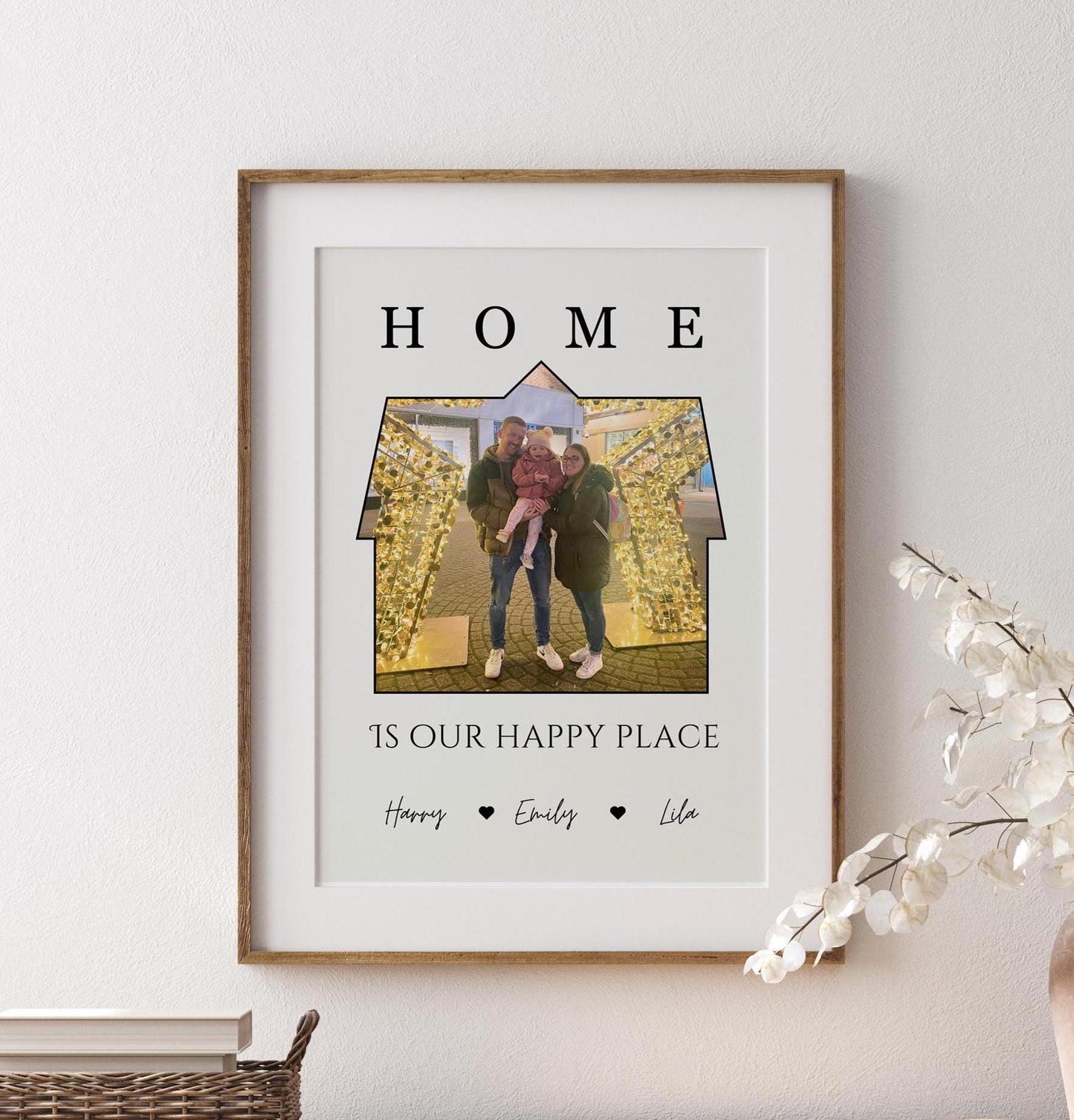 A framed print featuring a a photo set within the outline of a house. Above the image, the word &#39;HOME&#39; is written in bold, capital letters. Below the house-shaped frame, the phrase &#39;IS OUR HAPPY PLACE&#39; is printed in a stylish font. The bottom of the print includes the names &#39;Charlie, Sarah, Effie, Ralphie&#39; separated by small heart symbols. The frame is black, and the print has a clean, minimalist design.