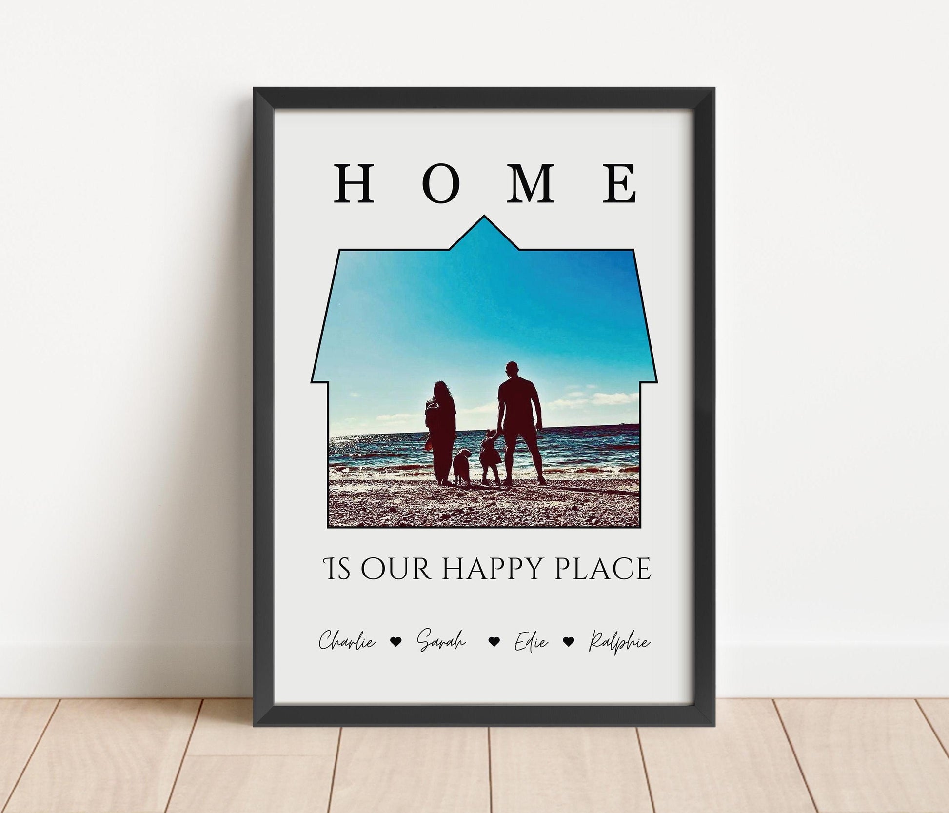A framed print featuring a a photo set within the outline of a house. Above the image, the word &#39;HOME&#39; is written in bold, capital letters. Below the house-shaped frame, the phrase &#39;IS OUR HAPPY PLACE&#39; is printed in a stylish font. The bottom of the print includes the names &#39;Charlie, Sarah, Effie, Ralphie&#39; separated by small heart symbols. The frame is black, and the print has a clean, minimalist design.