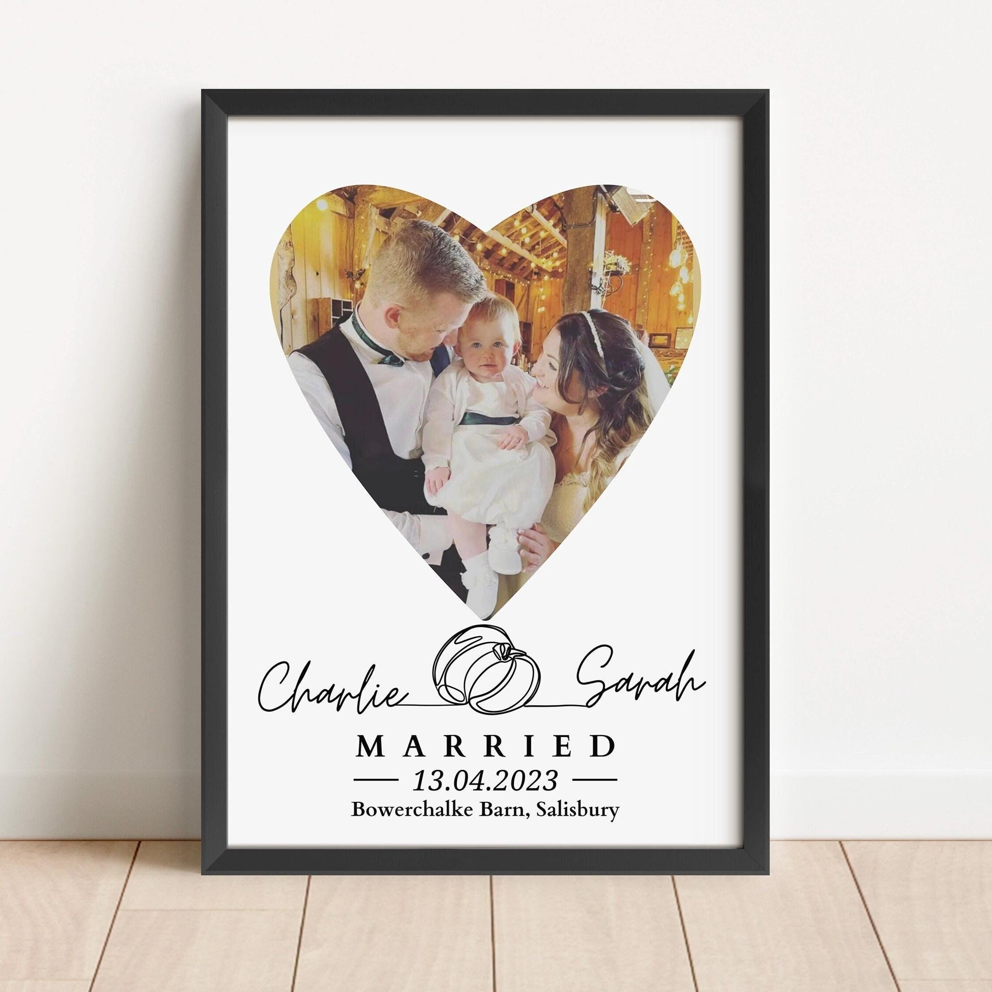 Framed wedding photo print featuring a couple with a child inside a heart-shaped photo design, personalized with names, wedding date, and venue.