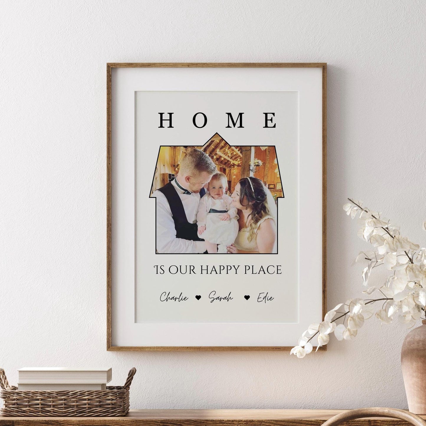 A framed print featuring a a photo set within the outline of a house. Above the image, the word &#39;HOME&#39; is written in bold, capital letters. Below the house-shaped frame, the phrase &#39;IS OUR HAPPY PLACE&#39; is printed in a stylish font. The bottom of the print includes the names &#39;Charlie, Sarah, Effie, Ralphie&#39; separated by small heart symbols. The frame is black, and the print has a clean, minimalist design.