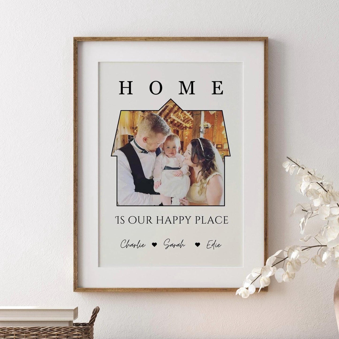 A framed print featuring a a photo set within the outline of a house. Above the image, the word &#39;HOME&#39; is written in bold, capital letters. Below the house-shaped frame, the phrase &#39;IS OUR HAPPY PLACE&#39; is printed in a stylish font. The bottom of the print includes the names &#39;Charlie, Sarah, Effie, Ralphie&#39; separated by small heart symbols. The frame is black, and the print has a clean, minimalist design.