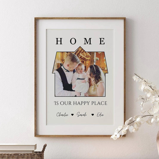 A framed print featuring a a photo set within the outline of a house. Above the image, the word &#39;HOME&#39; is written in bold, capital letters. Below the house-shaped frame, the phrase &#39;IS OUR HAPPY PLACE&#39; is printed in a stylish font. The bottom of the print includes the names &#39;Charlie, Sarah, Effie, Ralphie&#39; separated by small heart symbols. The frame is black, and the print has a clean, minimalist design.